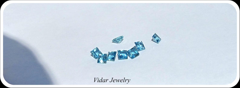 May include: A collection of light blue square-shaped gemstones on a white surface. The text "Vidar Jewelry" is printed below the gemstones.