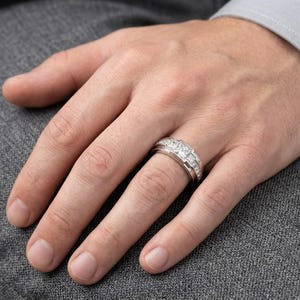 May include: A silver ring with three square-cut stones and smaller accent stones is worn on a finger. The ring is set against a gray background, with the hand and part of a gray suit visible.