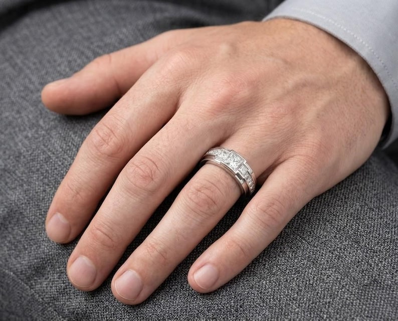May include: A silver ring with three square-cut stones and smaller accent stones is worn on a finger. The ring is set against a gray background, with the hand and part of a gray suit visible.