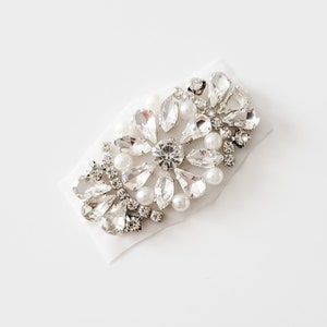 May include: A silver and white rhinestone and pearl embellishment for clothing or accessories.