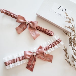 May include: Two dusty rose satin garters with white lace trim and central rhinestone accents. One garter features the embroidered text "You're Next!" on the ribbon tails, while the other displays "Mrs. Piskorski" and "February 10, 2024" in white thread.