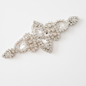May include: A silver rhinestone embellishment with a teardrop shape. The embellishment is made of small, clear rhinestones set in a silver metal frame.