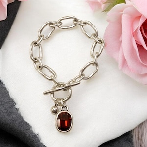 May include: Silver-toned chain bracelet with an oval, dark red gemstone pendant. The bracelet features a toggle clasp and a small, round charm. The jewelry is displayed on a white and black fabric, with pink roses in the background.