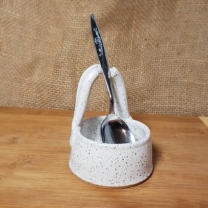 May include: White ceramic spoon rest with black speckles. The rest has a handle that holds a spoon upright.