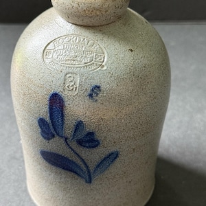 May include: A stoneware jug with a light gray base color and a dark blue floral design. The jug has a rounded body and a small, cylindrical neck. The text "ROCKDALE UNION STONEWARE" is stamped on the jug.