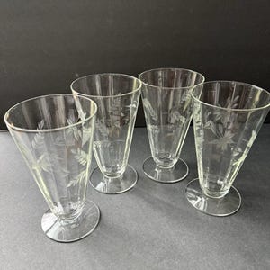 May include: Four clear, etched glass tumblers with a flared shape and round bases. Each glass features a delicate botanical design. The glasses are arranged on a dark surface, showcasing their elegant simplicity.