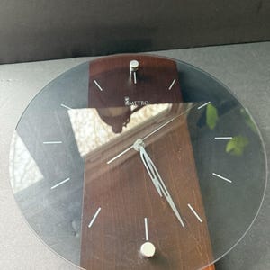 May include: A modern desk clock with a clear, circular face and silver hands. The clock features a dark wood base and a rectangular wooden support. The clock face has white markers and the brand name "METRO" is visible.
