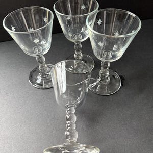 May include: Four clear glass cordial glasses with starburst designs. Each glass has a rounded bowl, a decorative stem, and a circular base. The starburst pattern is etched around the rim of each glass. The glasses are arranged on a dark surface.