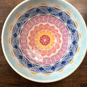 May include: A round ceramic bowl featuring a vibrant, hand-painted mandala design. The interior displays concentric circles of pink, orange, and deep blue, accented by white geometric lines and small yellow semi-circles along the rim.