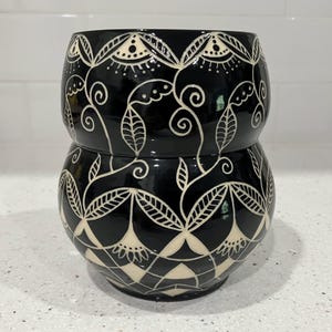 May include: A glossy black ceramic vessel with a rounded, bulbous shape and a cinched middle. The surface is decorated with intricate, hand-carved white line patterns featuring stylized floral motifs, swirling vines, and geometric leaf designs.
