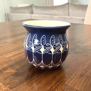 May include: A dark blue ceramic vase with a white floral pattern. The vase has a wide opening and a cream-colored interior. The design features stylized white flowers and geometric shapes. The vase is sitting on a wooden table.