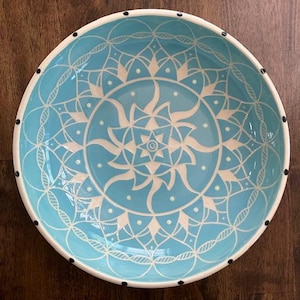 May include: A round, ceramic bowl with a light blue interior and a white, intricate mandala design. The rim is white with black dots. The bowl is a decorative piece for serving or display.