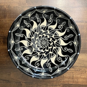 May include: A black ceramic decorative bowl with a white floral mandala design. The bowl has a glossy finish and a detailed pattern of swirling lines and leaf shapes. The rim is decorated with small white dots.