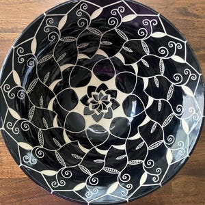 May include: A round, black ceramic bowl with a glossy finish. The bowl features a symmetrical design of cream-colored geometric shapes, leaves, and swirls. The center of the bowl has a floral design. The bowl is likely used for serving food or as a decorative piece.