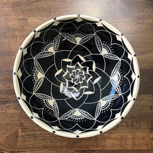 May include: A round ceramic bowl featuring a high-contrast black and white design. The interior surface is decorated with a symmetrical, intricate floral mandala pattern consisting of white lines on a black background, with a cream-colored rim.