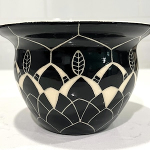 May include: A glossy black ceramic planter featuring a wide, flared rim and a white hand-painted geometric pattern. The design consists of repeating petal-like shapes and vertical leaf motifs that wrap around the exterior of the rounded bowl.