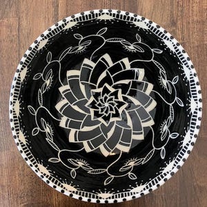 May include: A black and white ceramic bowl with a floral design. The bowl's interior features a stylized flower pattern in shades of gray and white. The rim is decorated with a repeating pattern of dots and lines, and white floral elements.
