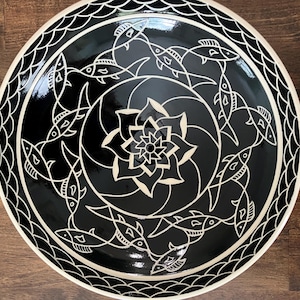 May include: A round ceramic plate featuring a glossy black glaze with white line art. The center displays a stylized floral motif surrounded by a circular pattern of swimming fish. The outer rim is decorated with a repeating wave-like pattern.