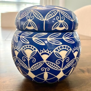 May include: A blue ceramic jar with a lid, adorned with a white floral and geometric pattern. The jar has a rounded shape and sits on a wooden surface. The design is symmetrical and detailed.