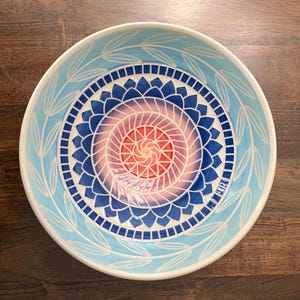 May include: A round ceramic bowl featuring a central red and orange spiral pattern surrounded by concentric rings of blue petals and small squares. The outer rim is light blue with a white leaf vine pattern painted along the interior edge.
