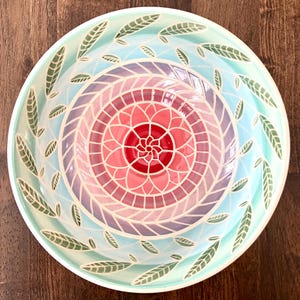 May include: A round ceramic bowl featuring a central red flower motif surrounded by concentric rings of pink and purple geometric patterns. The outer rim displays a light blue background decorated with a repeating pattern of green leaves.