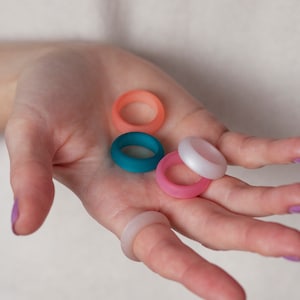 Könnte beinhalten: Fünf Silikonringe in verschiedenen Farben: Türkis, Rosa, Weiß und zwei Orangetönen. Die Ringe liegen übereinander in der Handfläche.