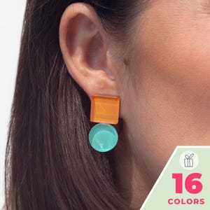 May include: A geometric earring featuring a translucent orange square stud positioned above a solid light turquoise circular drop. The earring is worn on an earlobe and is accompanied by a graphic overlay displaying the text 16 COLORS.