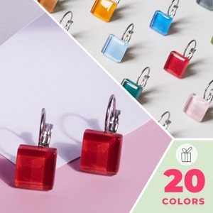 Puede incluir: Un par de pendientes cuadrados de vidrio rojo con aros de plata. Los pendientes están sobre una superficie rosa. La imagen también muestra una variedad de otros pendientes cuadrados de vidrio de colores con aros de plata. El texto "20 COLORS" está en la esquina inferior derecha de la imagen.