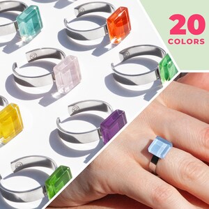 Könnte beinhalten: Ein Satz silberner Ringe mit bunten quadratischen Glassteinen. Die Ringe werden auf einer weißen Oberfläche präsentiert. Das Bild zeigt auch eine Hand, die einen Ring mit einem hellblauen Stein trägt. Der Text "20 COLORS" wird in der oberen rechten Ecke des Bildes angezeigt.