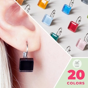 Puede incluir: Un par de pendientes cuadrados de cristal negro con aros plateados. Los pendientes se muestran en la oreja de una persona. La imagen también muestra una variedad de otros pendientes cuadrados de cristal de colores con aros plateados. El texto "20 COLORS" se muestra sobre un fondo verde.