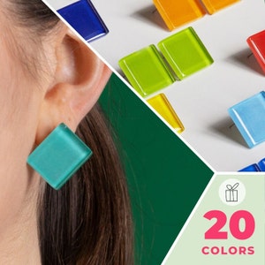 May include: A person wearing a turquoise square earring. The earring is made of glass and has a shiny finish. The background shows a variety of colorful square tiles, with the text "20 COLORS" in pink letters.