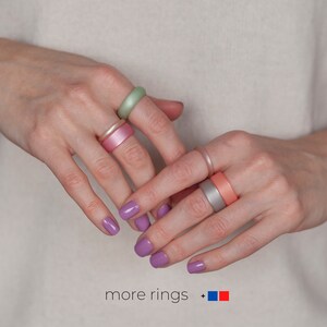 May include: A person's hands wearing a variety of silicone rings in different colors, including pink, green, and gray. The rings are stacked on the fingers and the person's nails are painted a light purple color. The text "more rings +" is visible at the bottom of the image.