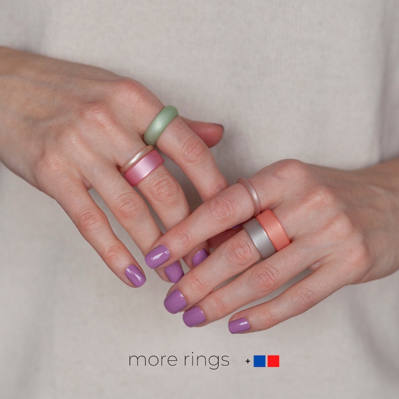May include: A person's hands wearing a variety of silicone rings in different colors, including pink, green, and gray. The rings are stacked on the fingers and the person's nails are painted a light purple color. The text "more rings +" is visible at the bottom of the image.