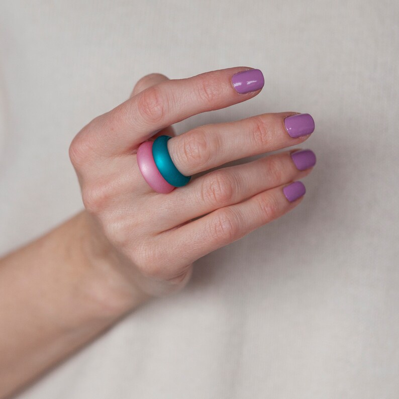 May include: A hand wearing two stacked rings, one teal and one pink. The nails are painted a light purple color.