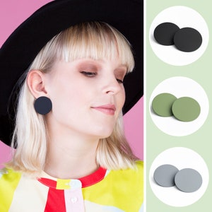 May include: A woman wearing a black hat and a colourful shirt with large black, green, and grey round earrings.