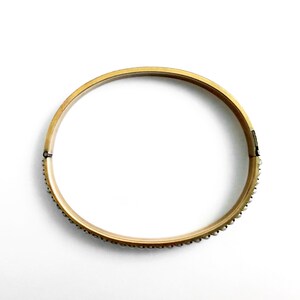 Puede incluir: Un brazalete dorado con cierre de bisagra. El brazalete est&aacute; adornado con peque&ntilde;os adornos redondos y espaciados a lo largo del borde exterior. La joya est&aacute; sobre un fondo blanco.