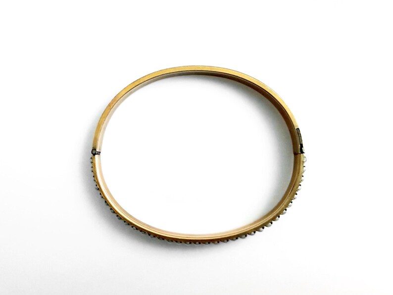 Puede incluir: Un brazalete dorado con cierre de bisagra. El brazalete est&aacute; adornado con peque&ntilde;os adornos redondos y espaciados a lo largo del borde exterior. La joya est&aacute; sobre un fondo blanco.