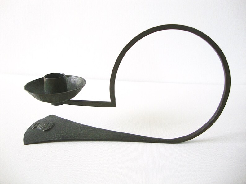 Puede incluir: Un candelero de metal negro con una base circular y un brazo curvo que sostiene una copa para velas. El candelero tiene una textura martillada y un dise&ntilde;o decorativo en la base.