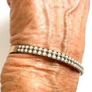 Puede incluir: Una pulsera dorada con dos filas de peque&ntilde;as perlas blancas. La pulsera se lleva en la mu&ntilde;eca. Las perlas est&aacute;n espaciadas uniformemente. La pulsera tiene un dise&ntilde;o sencillo y elegante.