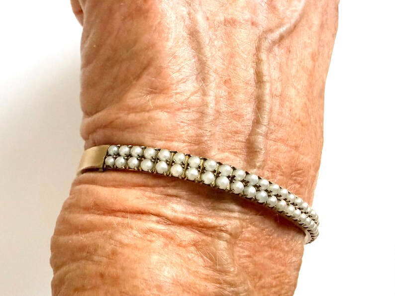Puede incluir: Una pulsera dorada con dos filas de peque&ntilde;as perlas blancas. La pulsera se lleva en la mu&ntilde;eca. Las perlas est&aacute;n espaciadas uniformemente. La pulsera tiene un dise&ntilde;o sencillo y elegante.