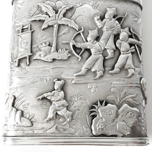May include: A silver rectangular box with an embossed design of people in a landscape. The people are dressed in traditional clothing and are holding bows and arrows. The landscape includes trees, mountains, and a river.