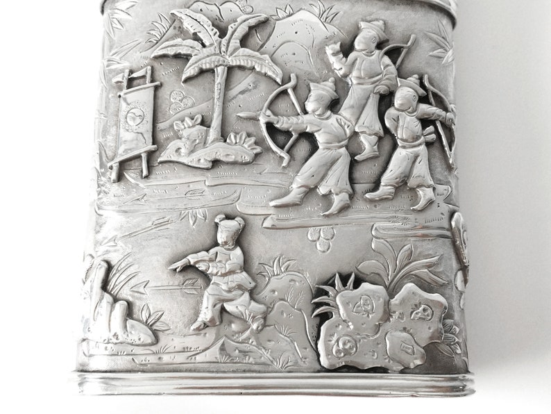 May include: A silver rectangular box with an embossed design of people in a landscape. The people are dressed in traditional clothing and are holding bows and arrows. The landscape includes trees, mountains, and a river.