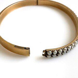 Puede incluir: Brazalete r&iacute;gido dorado con bisagras y dos filas de peque&ntilde;as perlas redondas grises. El brazalete est&aacute; abierto, revelando el mecanismo de cierre. El fondo es blanco.
