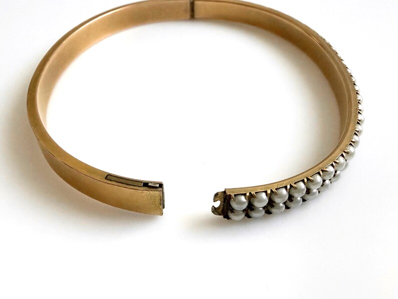 Puede incluir: Brazalete r&iacute;gido dorado con bisagras y dos filas de peque&ntilde;as perlas redondas grises. El brazalete est&aacute; abierto, revelando el mecanismo de cierre. El fondo es blanco.