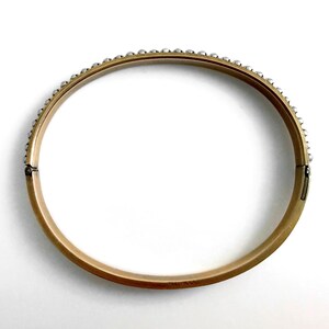 Puede incluir: Un brazalete dorado con una fila de peque&ntilde;as cuentas redondas de color plateado a lo largo del borde superior. El brazalete tiene un cierre de bisagra y est&aacute; sobre un fondo blanco. Una joya cl&aacute;sica.
