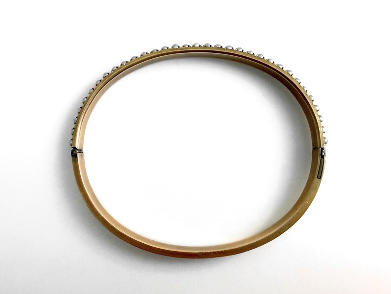 Puede incluir: Un brazalete dorado con una fila de peque&ntilde;as cuentas redondas de color plateado a lo largo del borde superior. El brazalete tiene un cierre de bisagra y est&aacute; sobre un fondo blanco. Una joya cl&aacute;sica.
