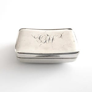 Puede incluir: Pequeña caja rectangular plateada con tapa curvada. La tapa está grabada con iniciales en escritura ornamental. La caja tiene un diseño con bisagras y un acabado pulido. Para joyas o pequeños recuerdos.