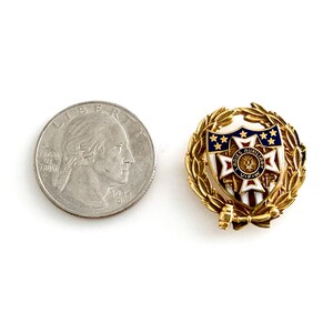 May include: A gold-toned pin with a shield design featuring blue stars and red stripes, surrounded by a laurel wreath. A quarter coin is next to the pin for size comparison.