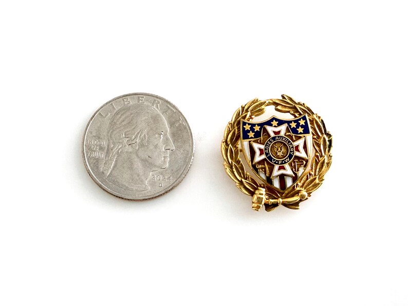 May include: A gold-toned pin with a shield design featuring blue stars and red stripes, surrounded by a laurel wreath. A quarter coin is next to the pin for size comparison.