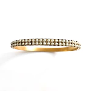 Puede incluir: Una pulsera dorada con dos filas de peque&ntilde;as perlas redondas de color blanco roto. Las perlas est&aacute;n muy juntas, creando una superficie texturizada. La pulsera est&aacute; sobre un fondo blanco.